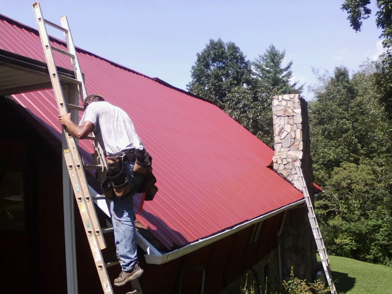 Roofer on a ladder beside a red metal roof and stone chimney — Soffit & Fascia Repair by Roofing Brothers in Rosemead
